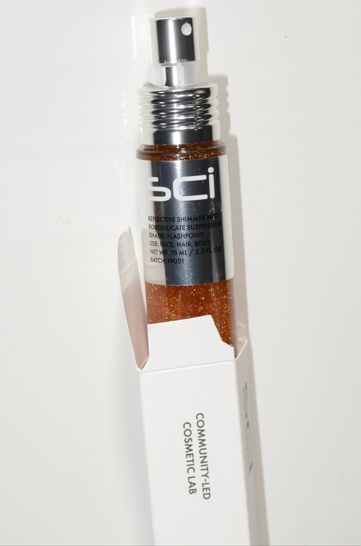 A bottle of SCI Cosmetics Bronzy Glow Bronzing Powder with a silver spray pump, shown partially inside its white cardboard packaging box against a plain white background.