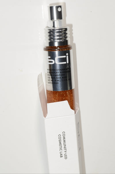 A bottle of SCI Cosmetics Bronzy Glow Bronzing Powder with a silver spray pump, shown partially inside its white cardboard packaging box against a plain white background.